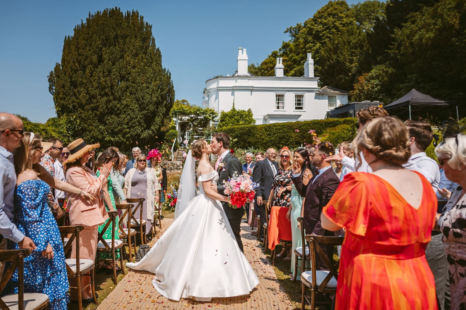 Confetti moment and kiss at Incredible wedding venue Remenham Court on hot summers day with blue sky above white house