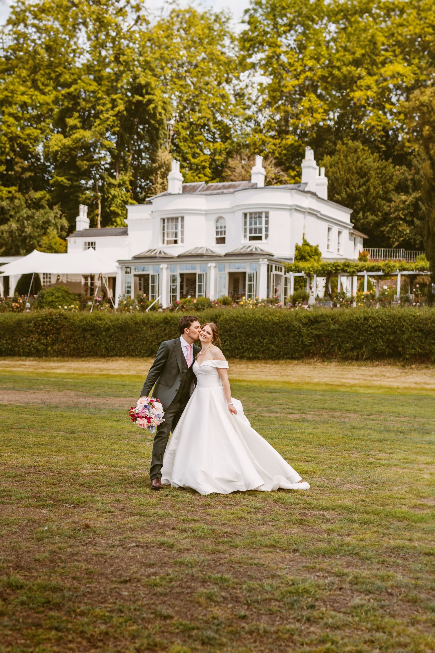 Beautiful bride and Groom laughing together walking across lawn at Remenham Court Henley on Thames pausing for kiss