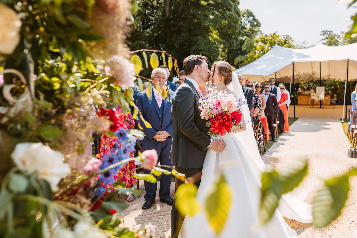 Bride arriving at top of aisle and greeting groom surrounded by bold colourful flowers summers day