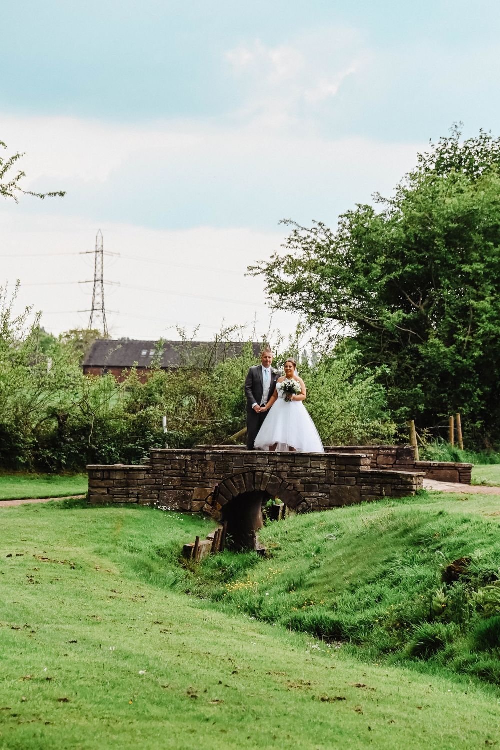 Wedding couple standing on rustic stone bridge in countryside setting.