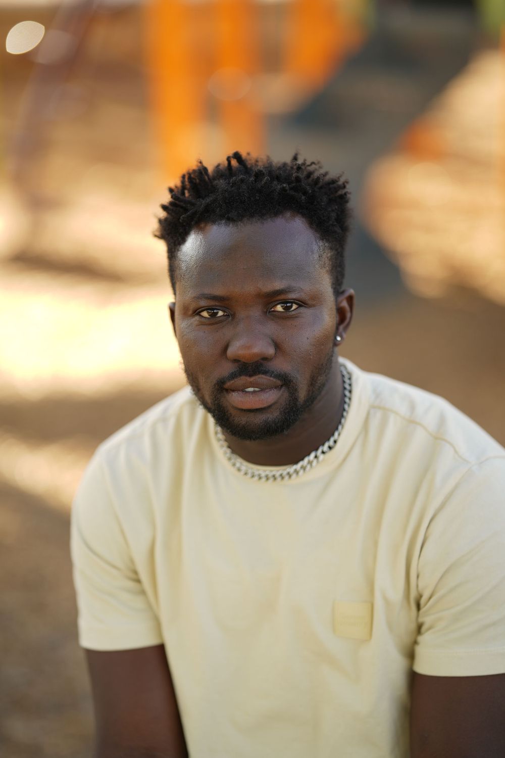 A person in a cream colored t-shirt with a chain necklace poses outdoors in warm lighting.