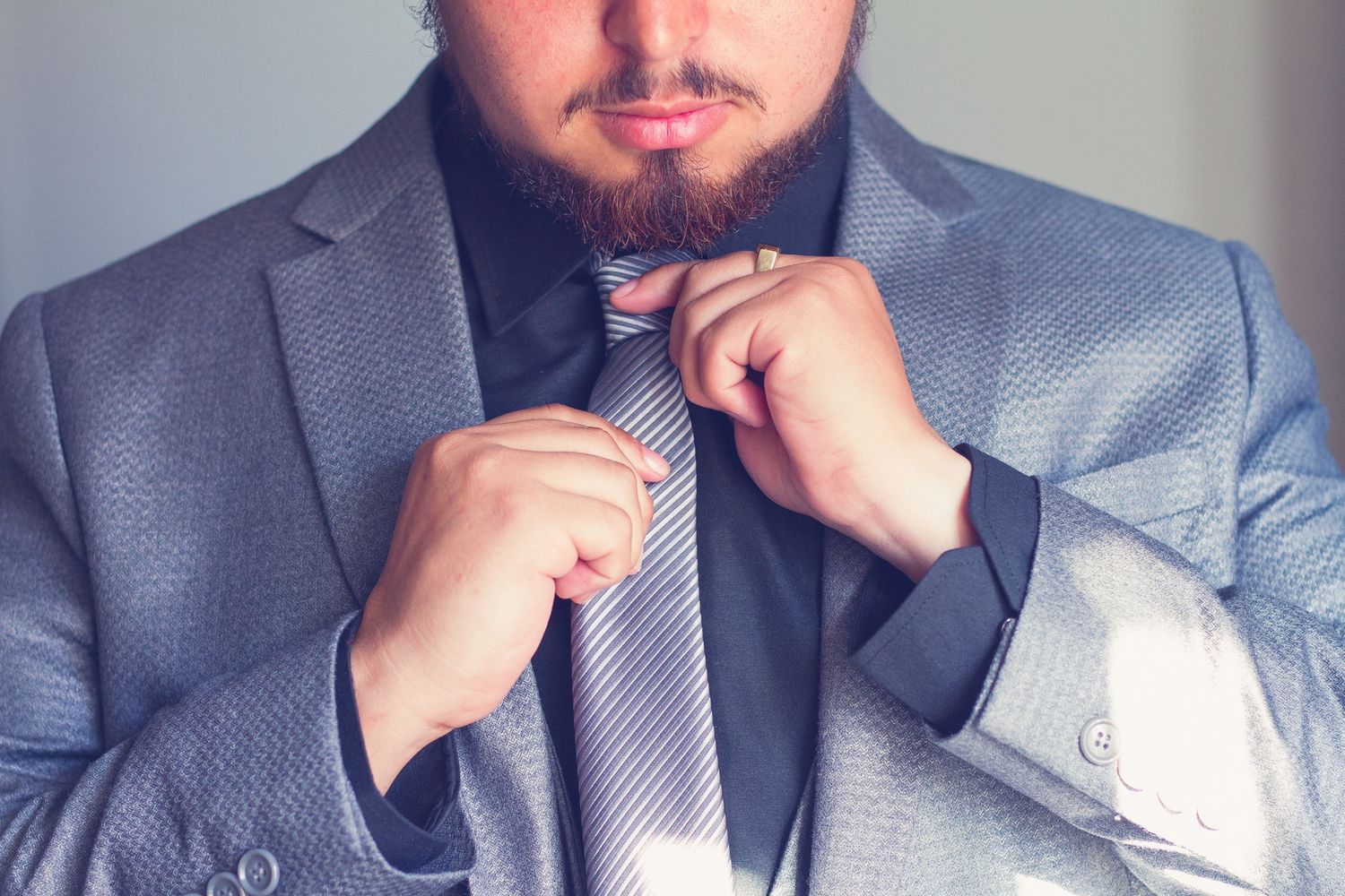 Bearded man in a gray suit adjusts his striped tie while getting ready for a formal event.