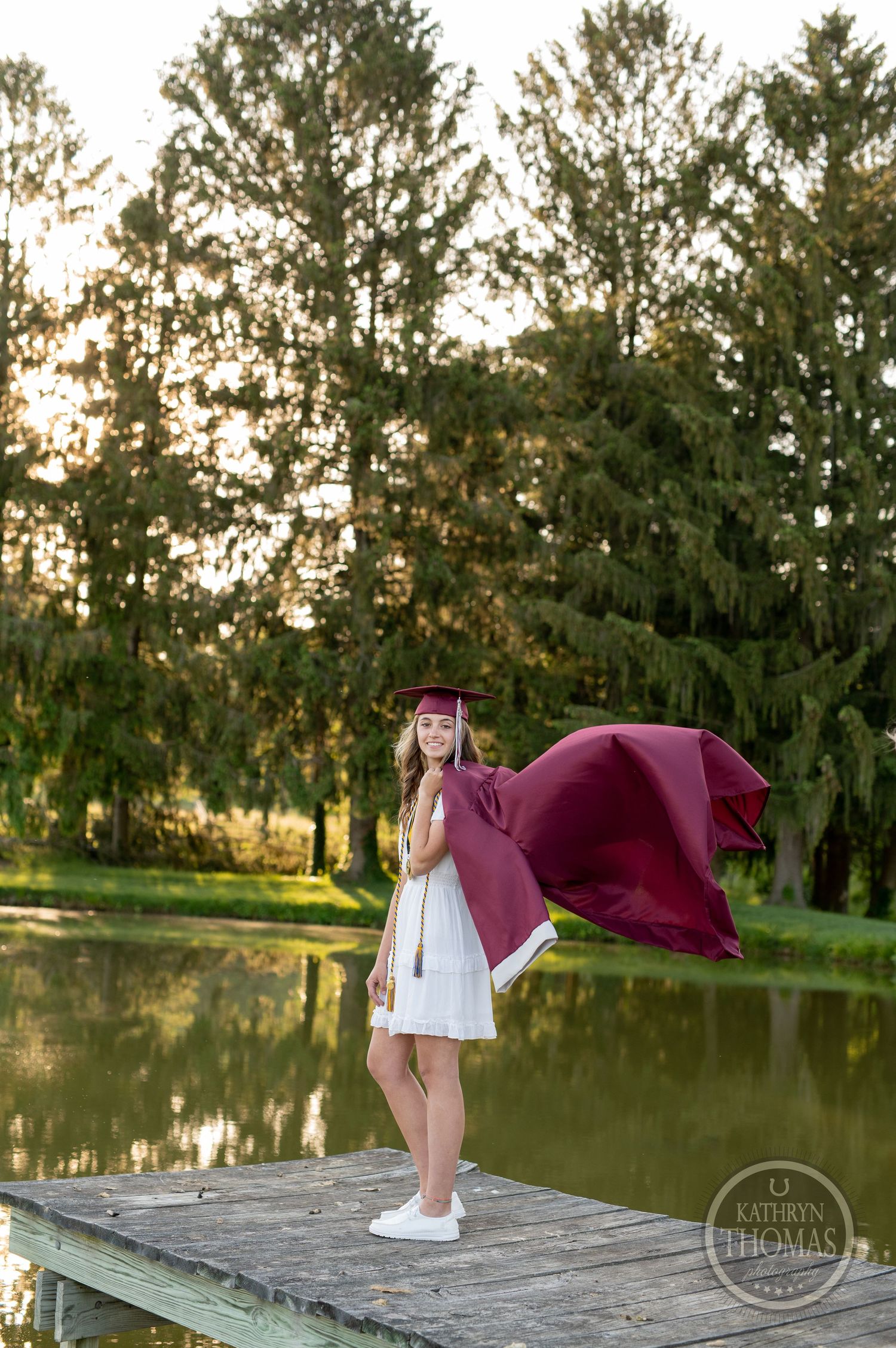 Graduate's burgundy gown flows in breeze while standing on dock by pond at sunset.