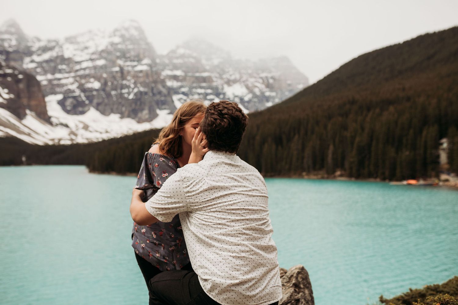 A romantic embrace on rocky shores with turquoise waters and mountain peaks in the distance.