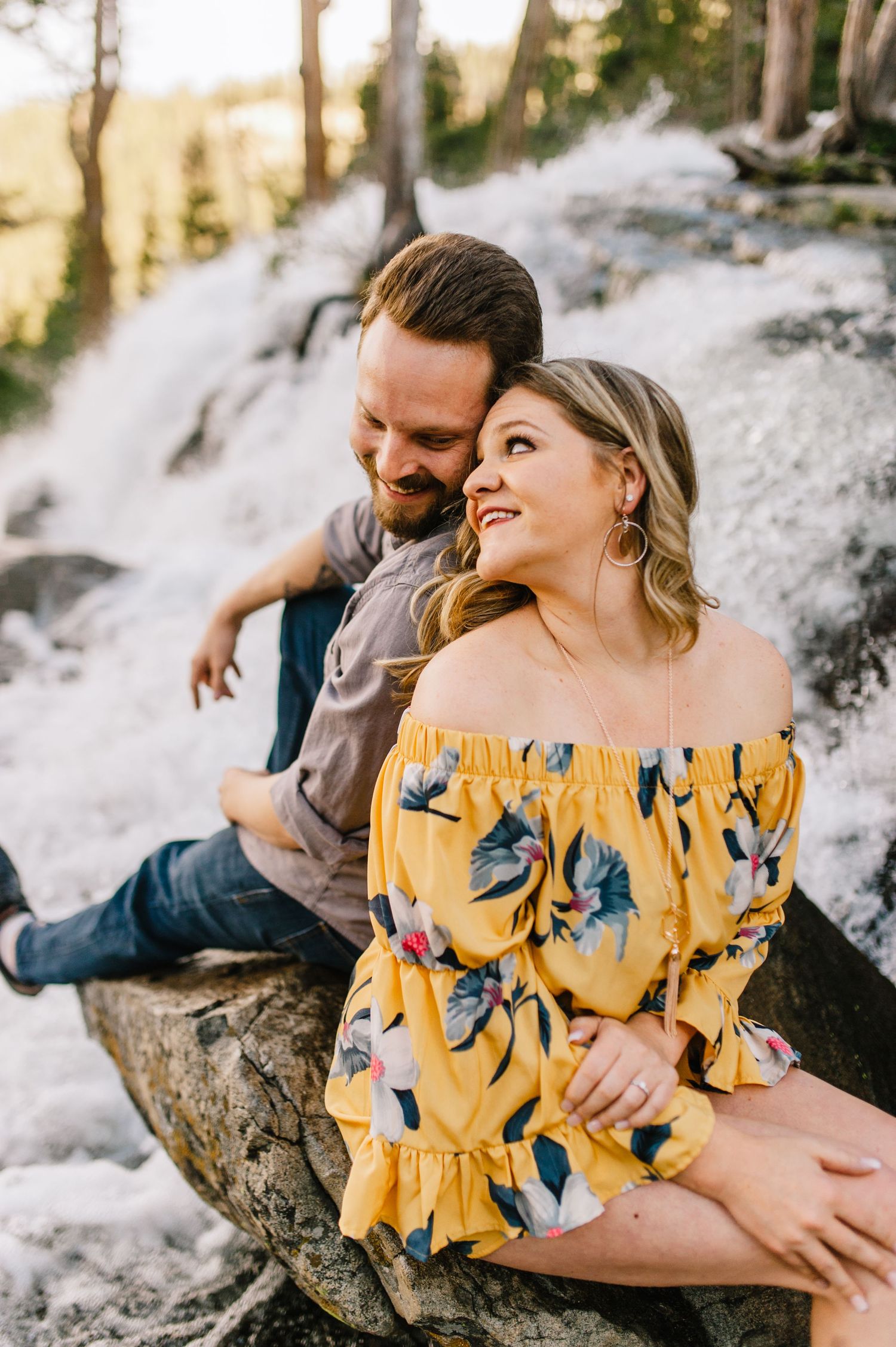 Couple sitting intimately together on a rock by a waterfall with woman wearing yellow floral off-shoulder dress.