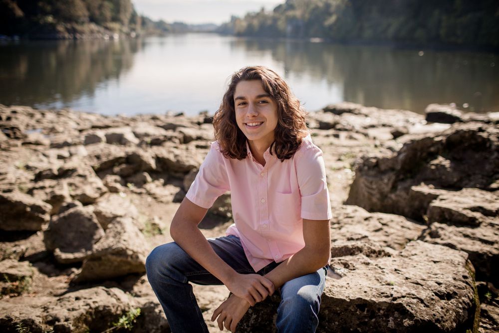 Teenage boy in pink polo shirt sitting on rocks near a peaceful river in autumn.