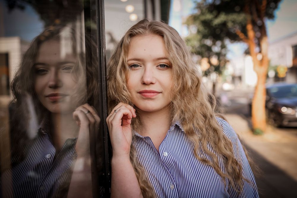 Young blonde woman leaning against a glass window with her reflection visible in an urban portrait.