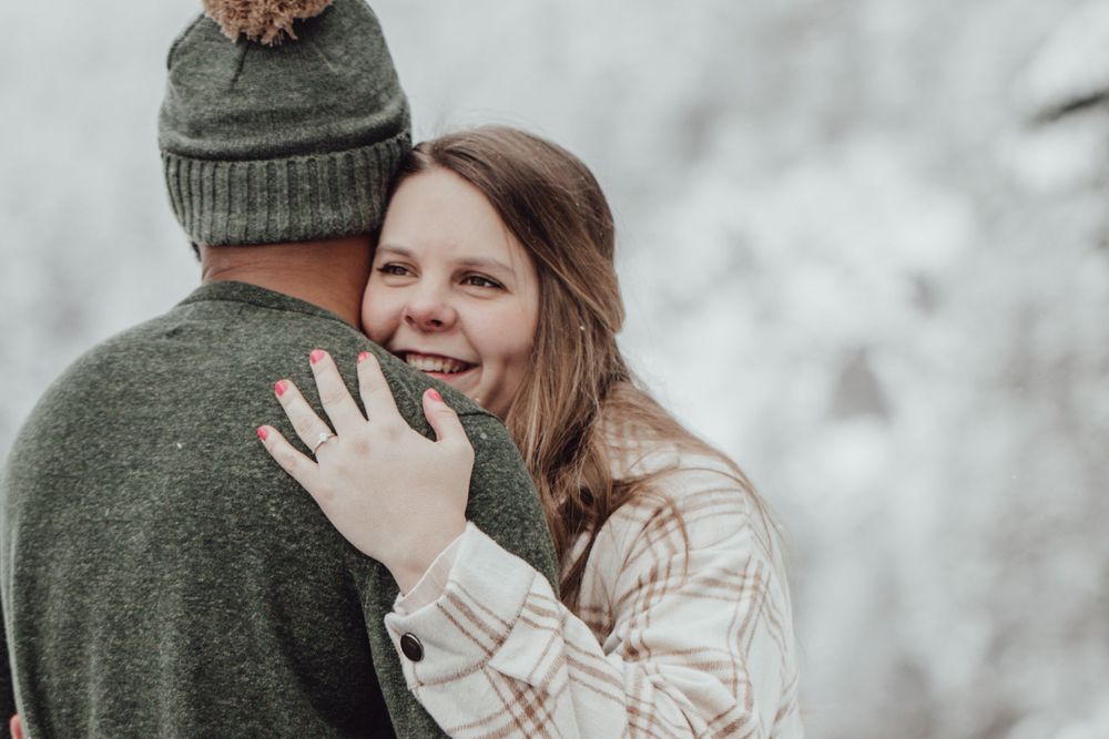 A couple hugs while the woman shows off her ring, on a snowy pier during an engagement session with Pamela Clark