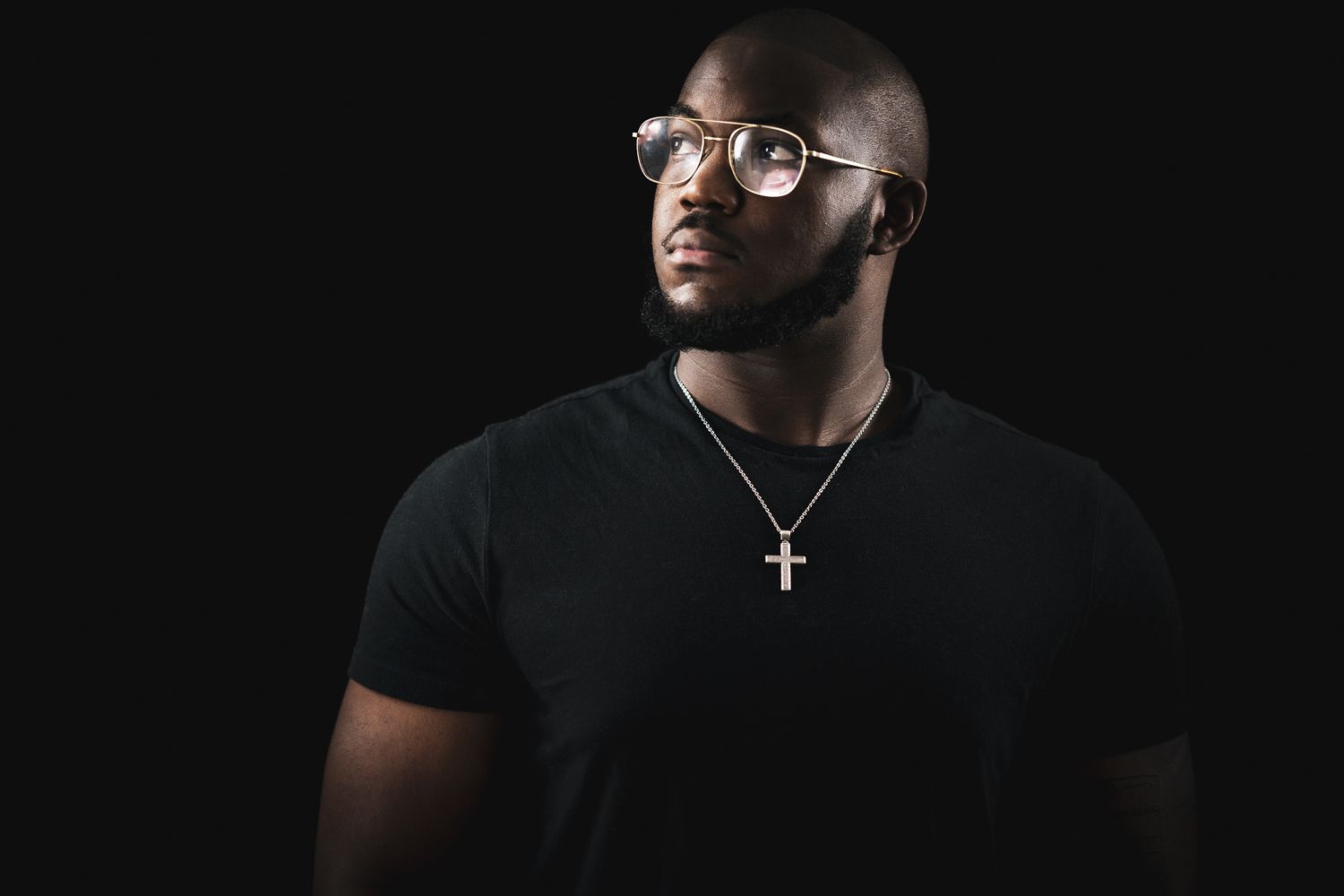 A studio portrait of a person in glasses wearing a black t-shirt and cross necklace against dark background.