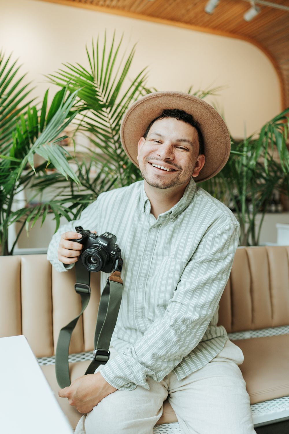 Photographer in casual attire and hat sits on yellow couch with camera surrounded by tropical plants.