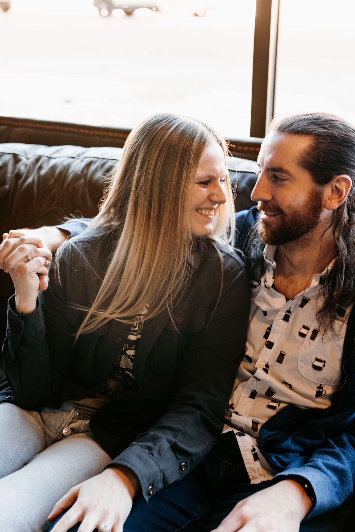 A couple shares a candid laughing moment while sitting on a leather couch by a window.