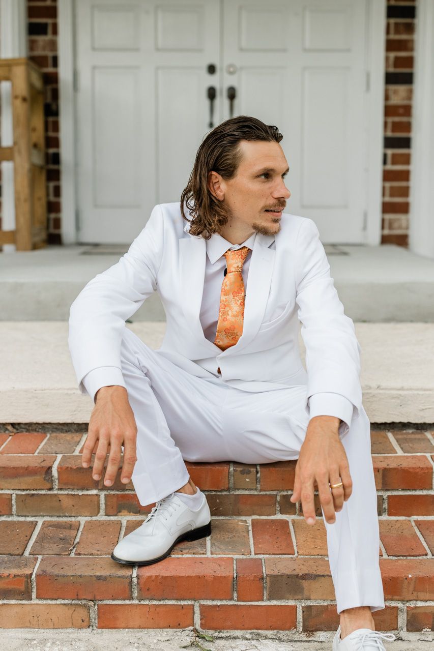 A person in all white attire with orange tie sits casually on brick steps.