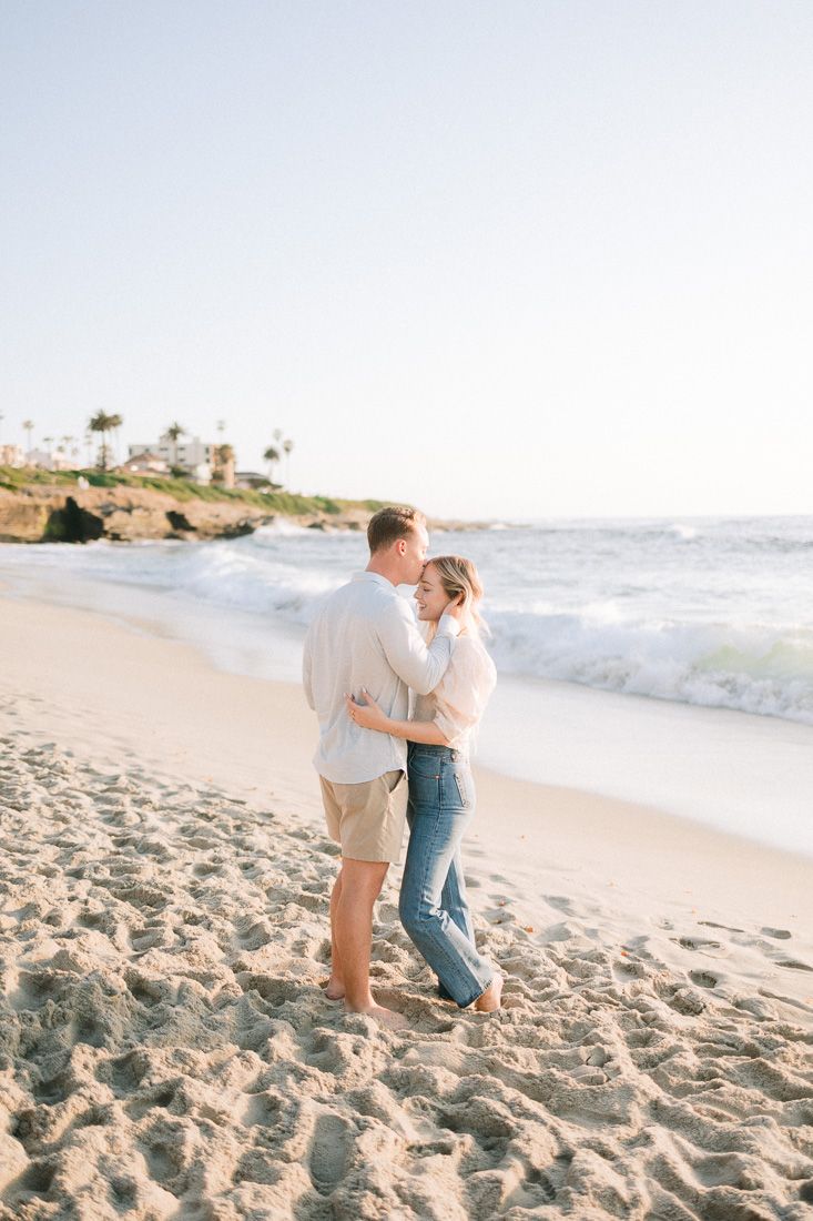 A romantic couple embraces while standing on a sandy beach with palm trees and ocean waves in the background.