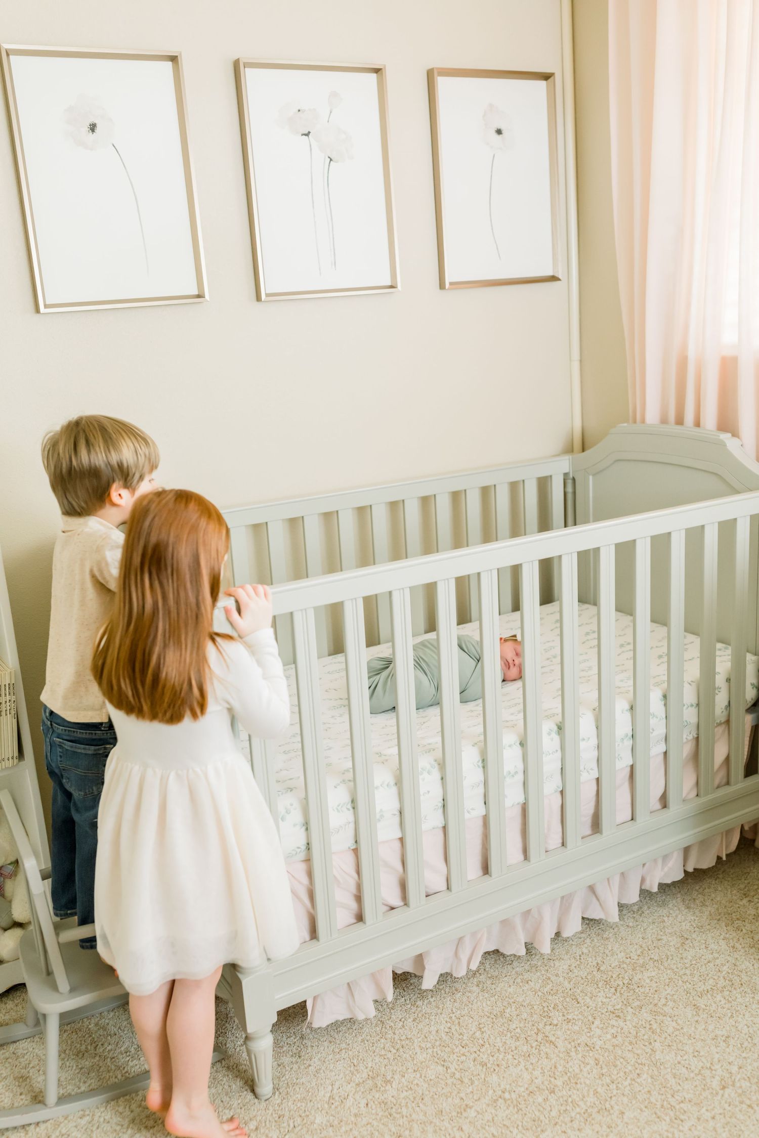 Two children peek into a gray crib with pink bedding beneath minimalist floral artwork.