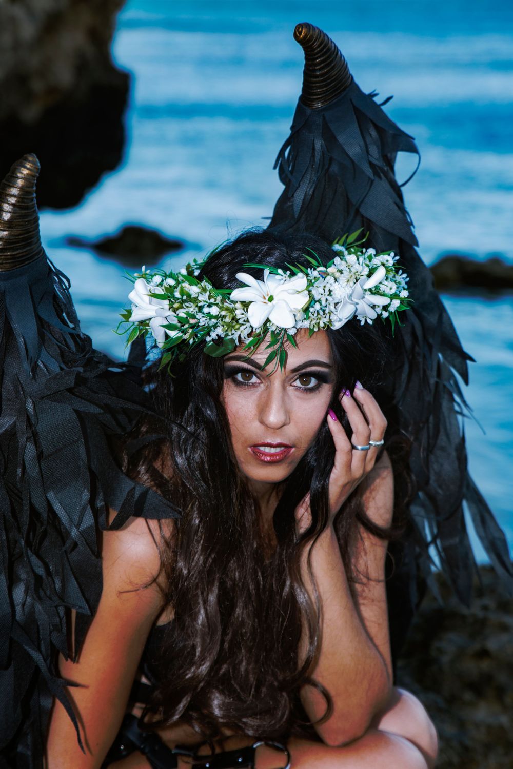 Beach portrait with dramatic black wings and white floral crown against blue ocean backdrop.