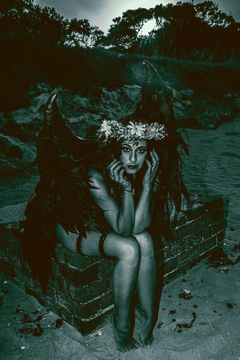 Moody nighttime portrait with black wings and flower crown in dramatic blue lighting.