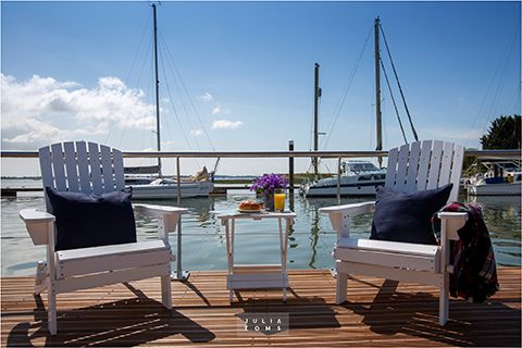 White Adirondack chairs on wooden dock overlooking peaceful marina with sailboats.