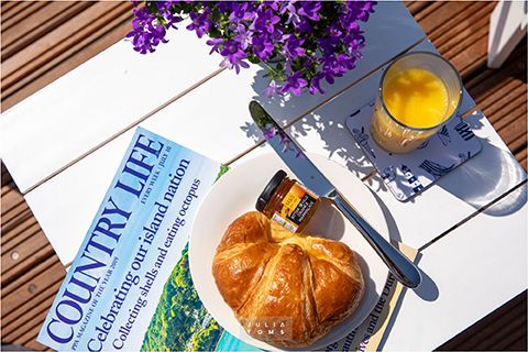 Breakfast setting with a croissant, magazine and purple flowers on a wooden table in sunlight.
