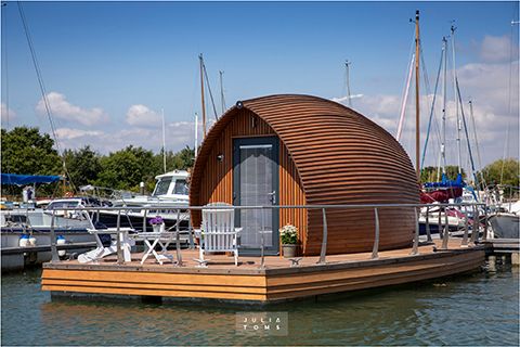 Wooden arch-shaped houseboat with deck moored at a marina among sailing vessels.