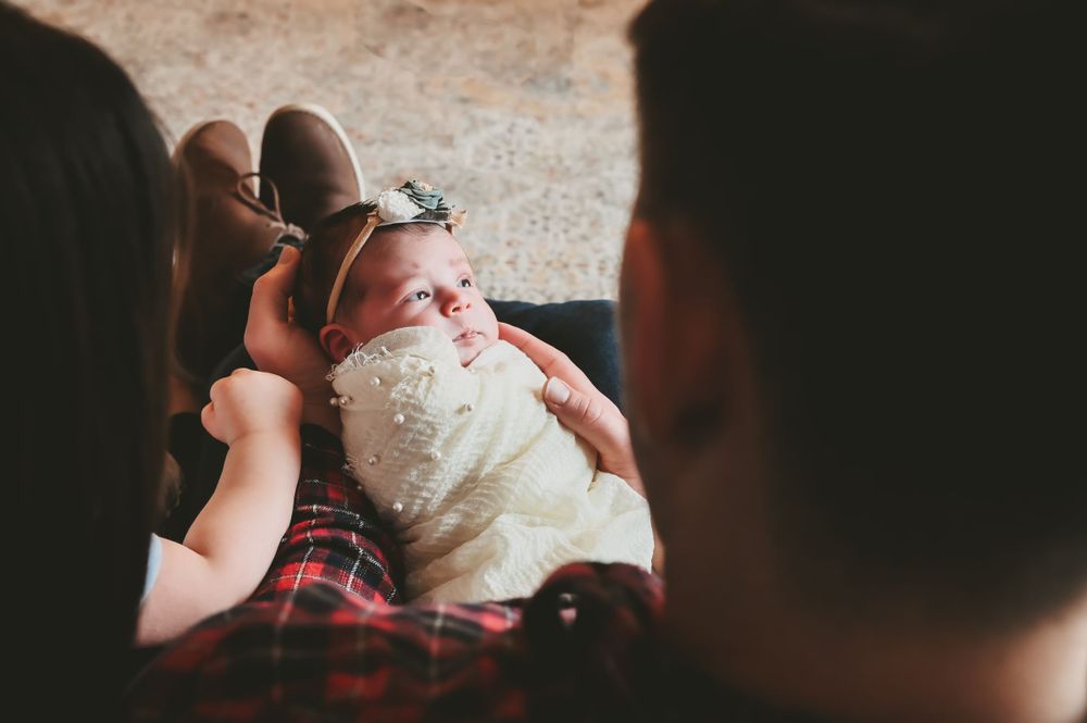 Newborn baby in white wrap being held by parents in casual attire.