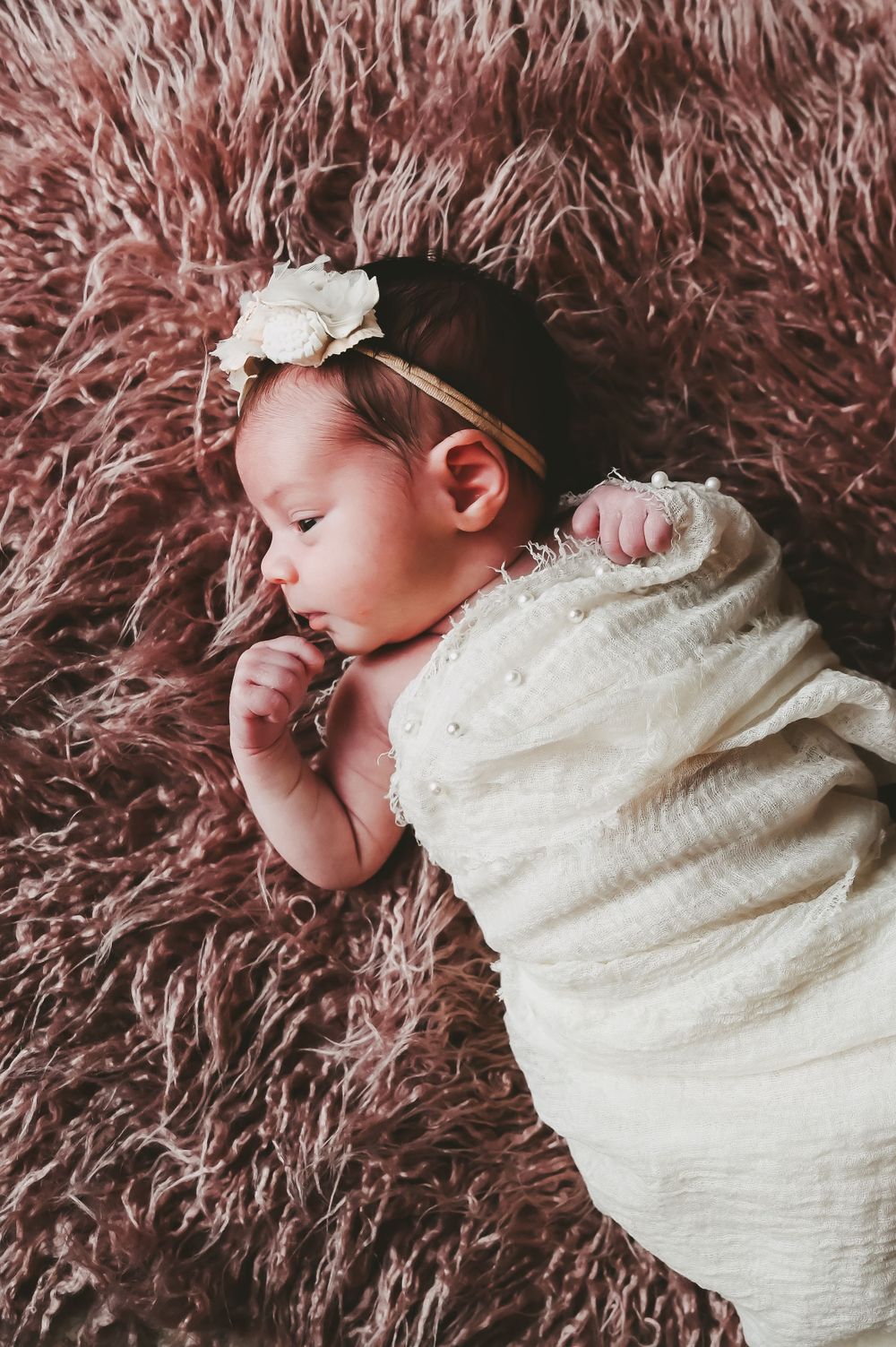 Peaceful newborn baby wearing white bow headband sleeping on pink shag carpet.