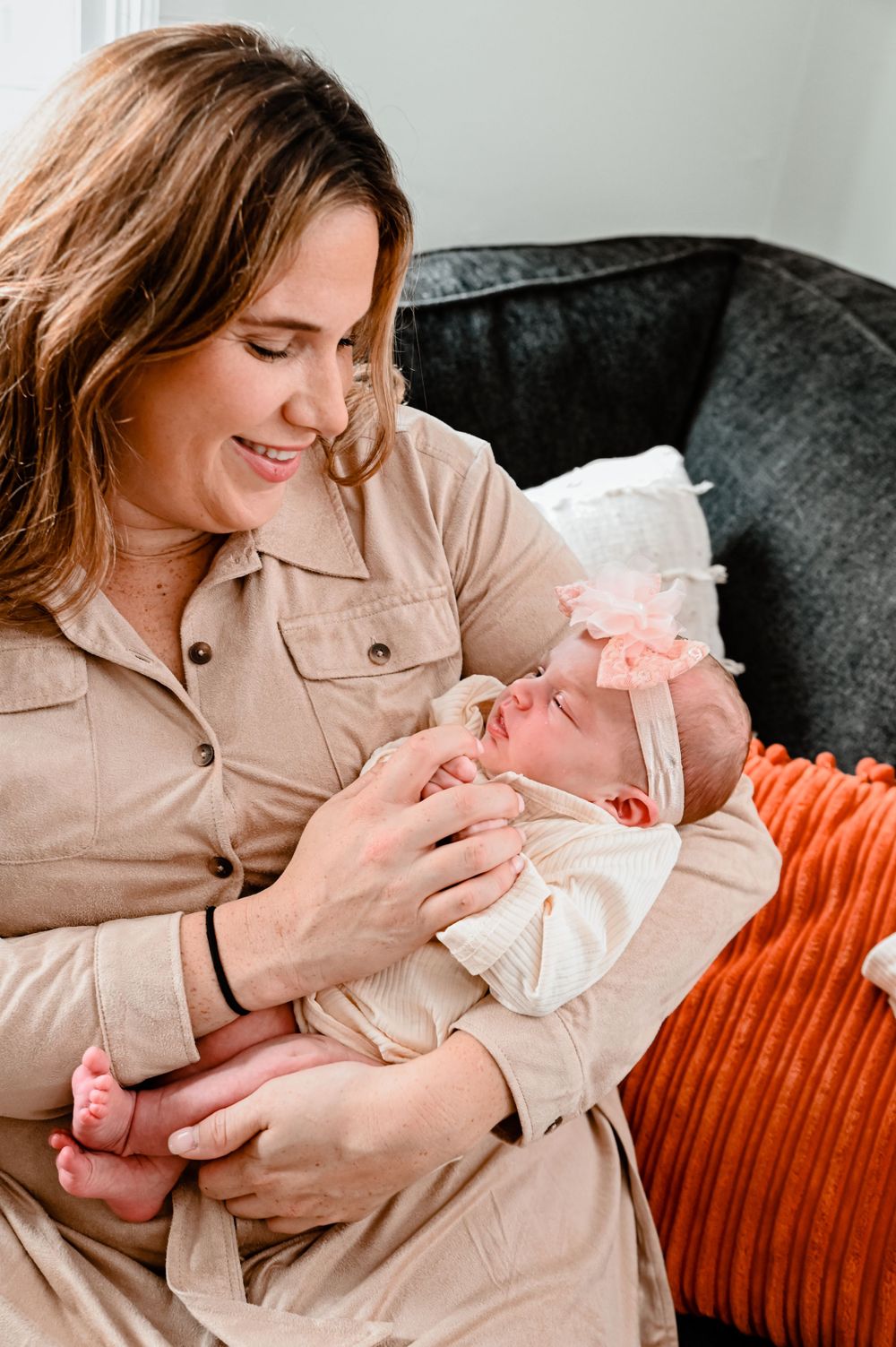 Mother in beige jacket snuggling with newborn baby on gray couch.
