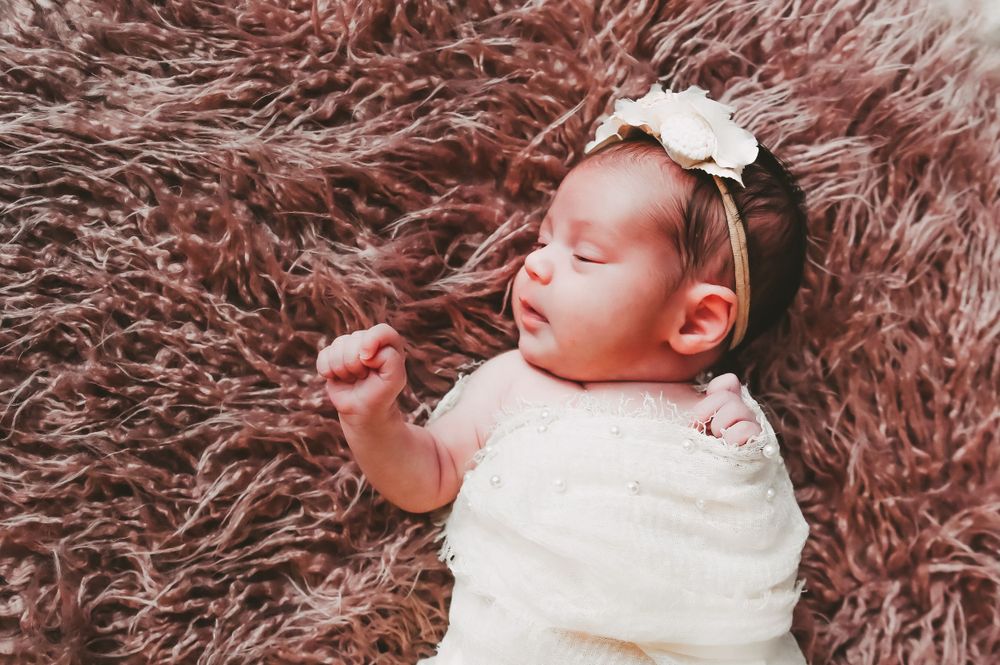 Sleeping newborn baby with white bow headband lying on pink fluffy rug.