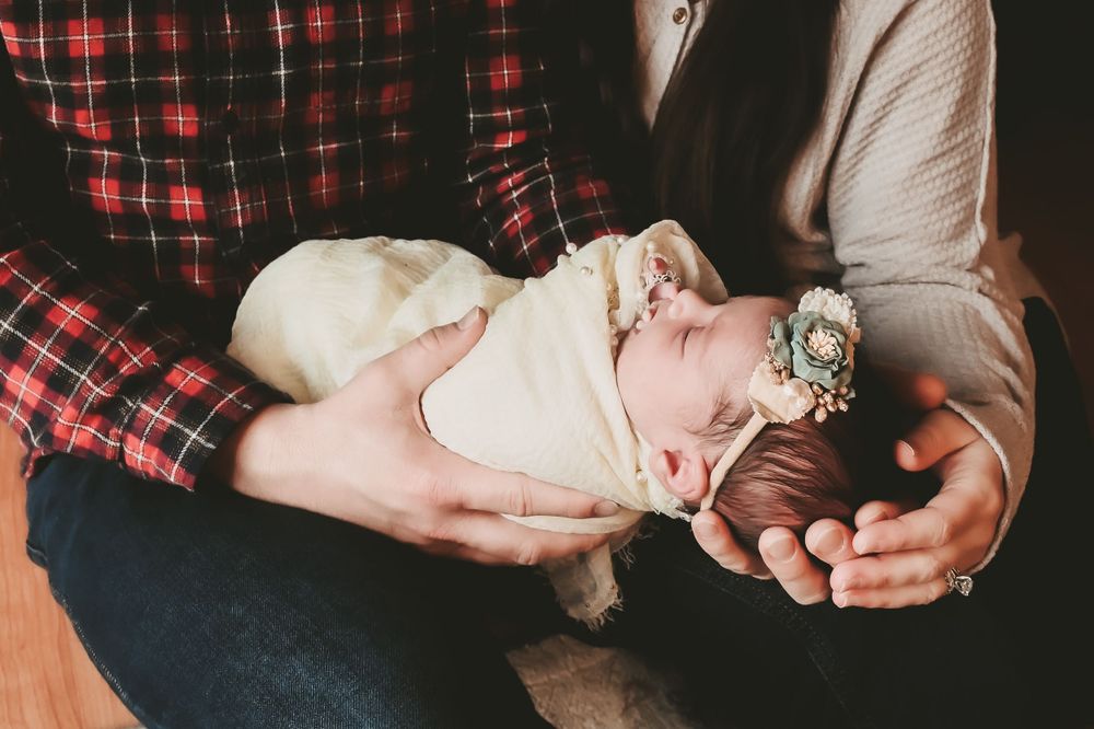 Newborn baby in white wrap with floral headband cradled in arms wearing plaid.