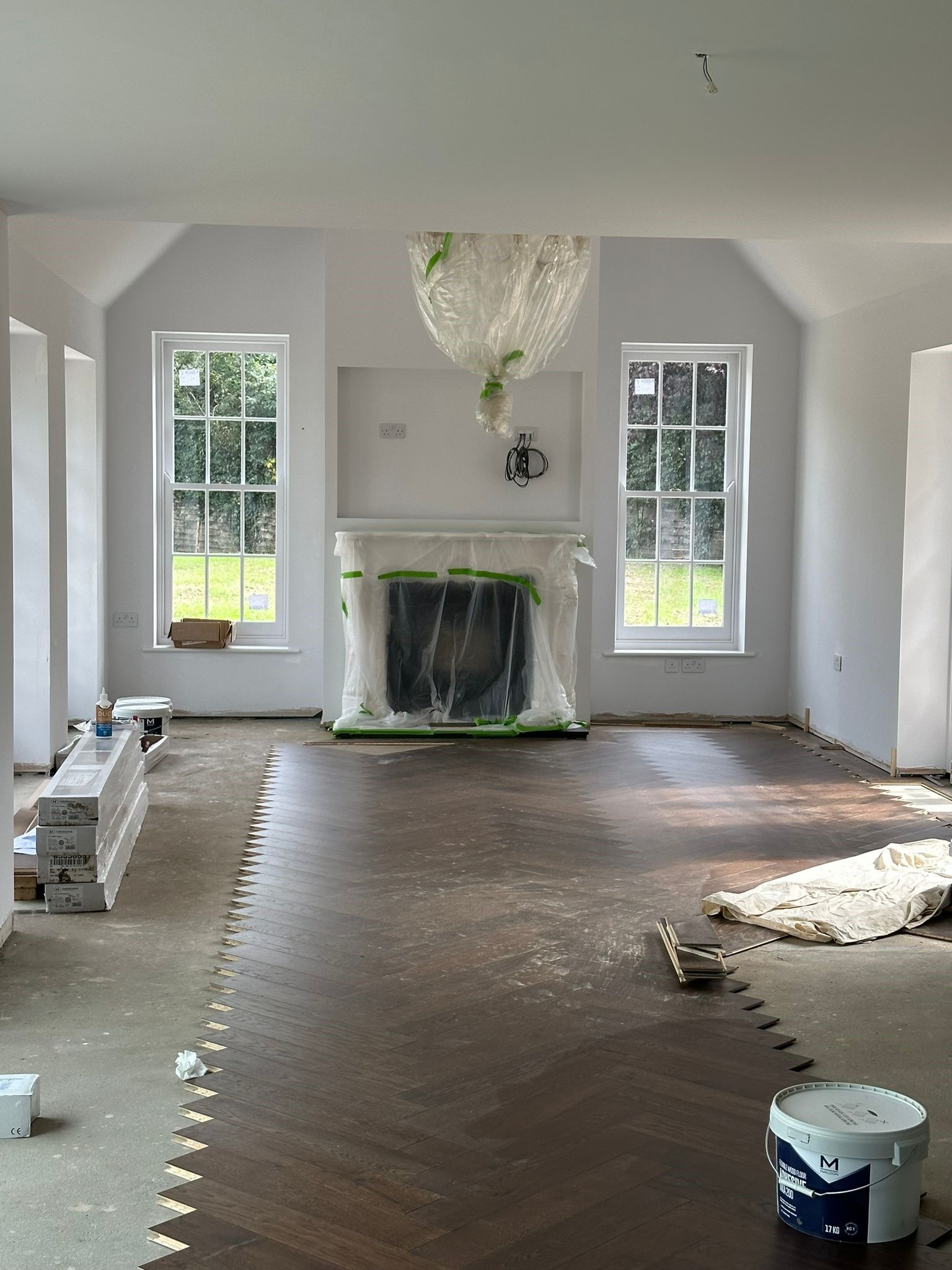 Under-construction living room with herringbone flooring, symmetrical windows, and central fireplace.