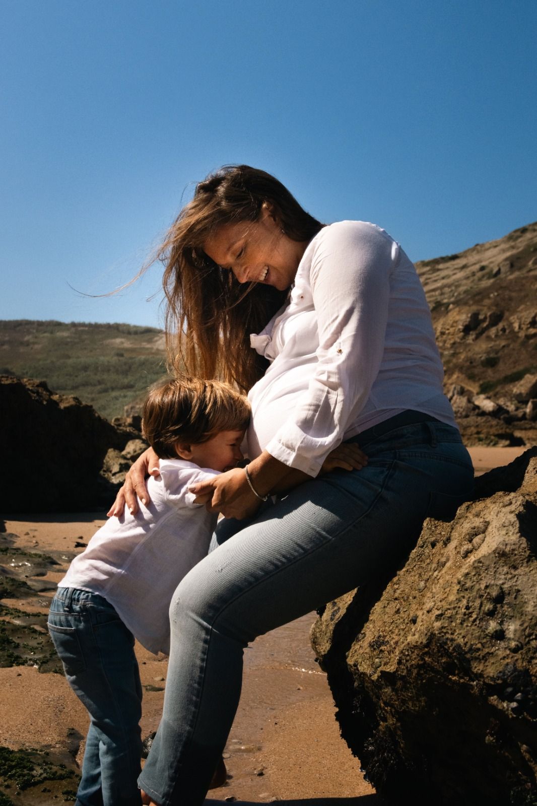 Two people sitting together on rocky coastal terrain under bright blue sky.