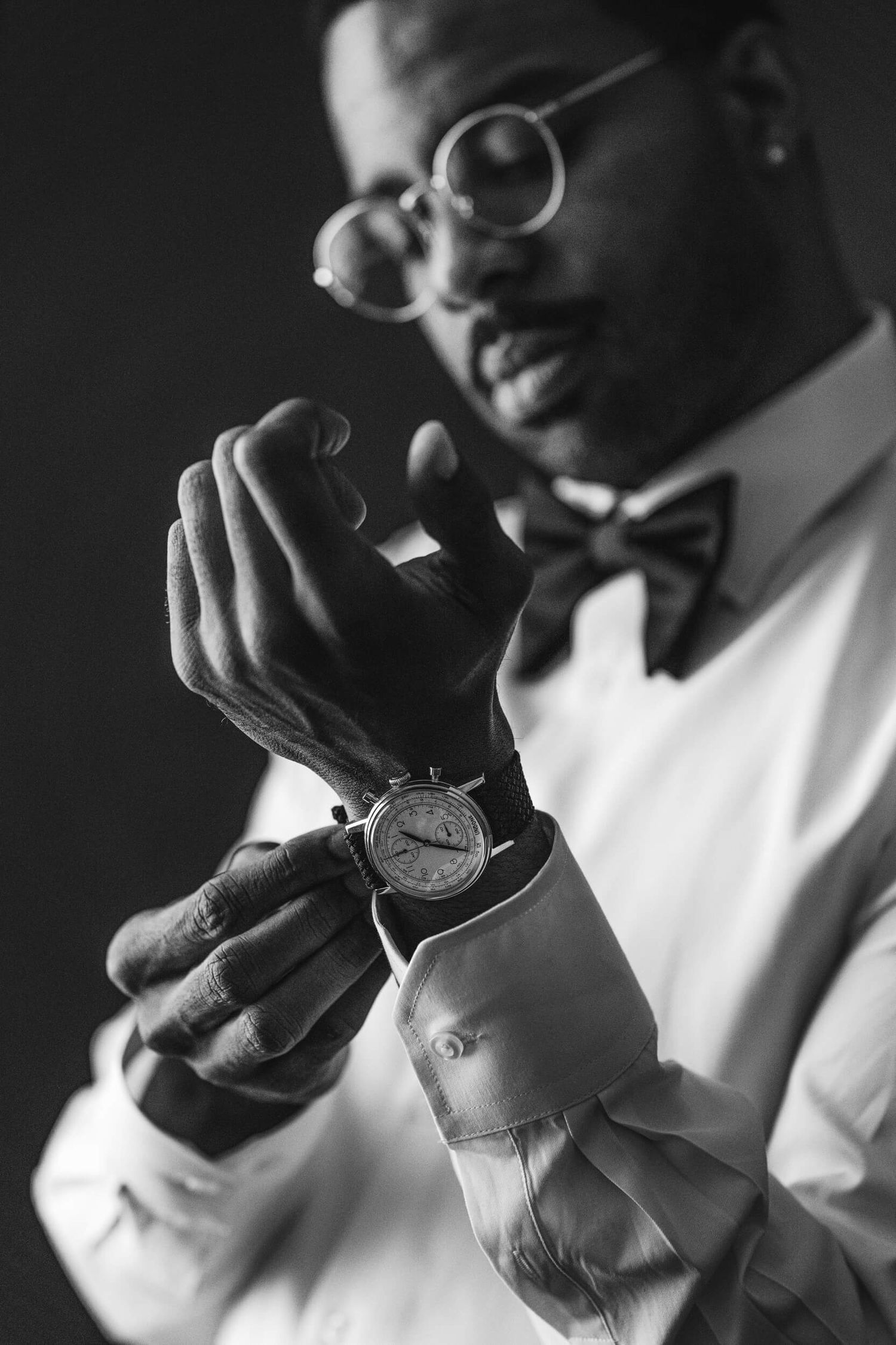 Black and white portrait of a groom checking his watch while wearing a bowtie and glasses.