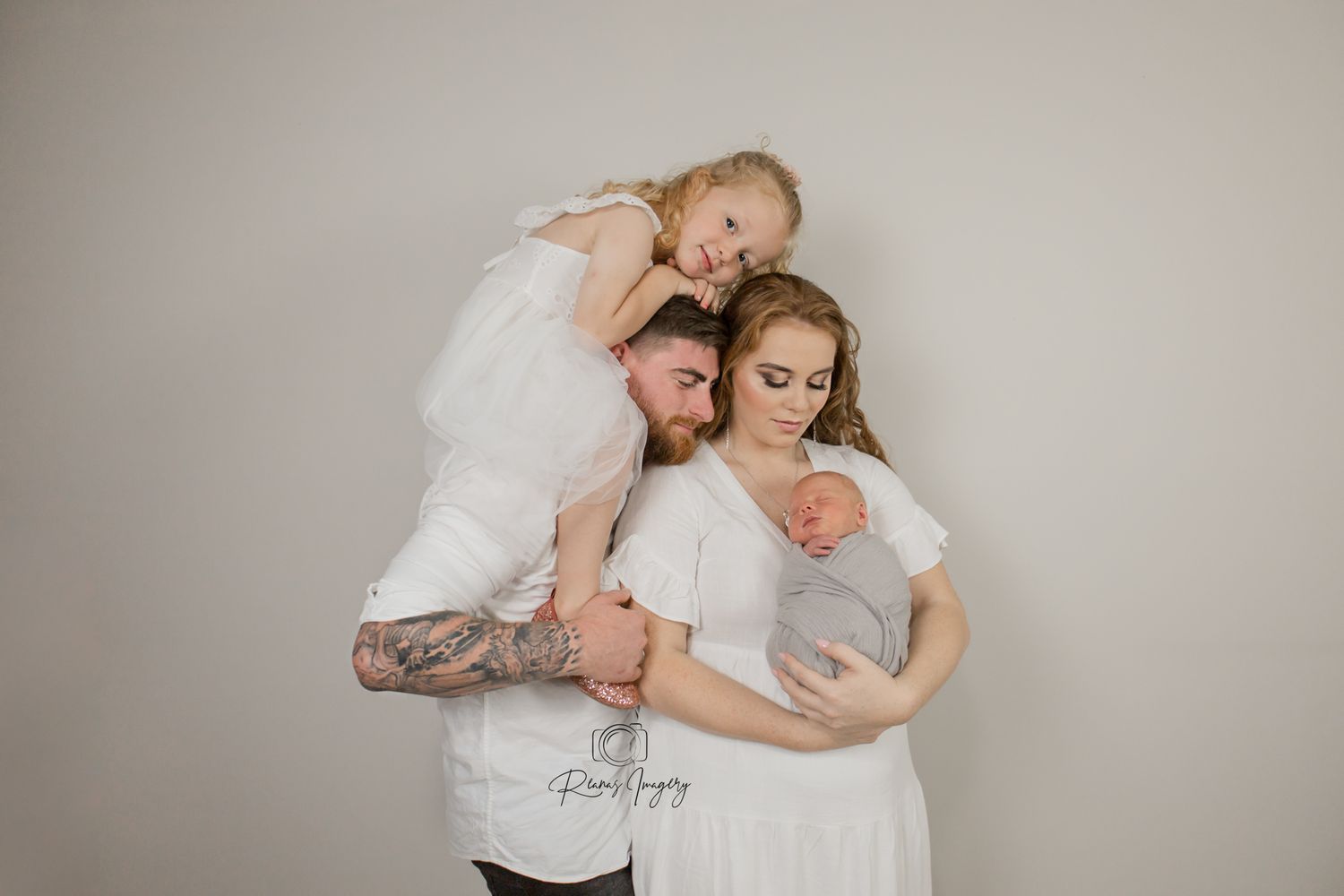 Family studio portrait with tattooed dad holding toddler girl while mom cradles newborn baby.