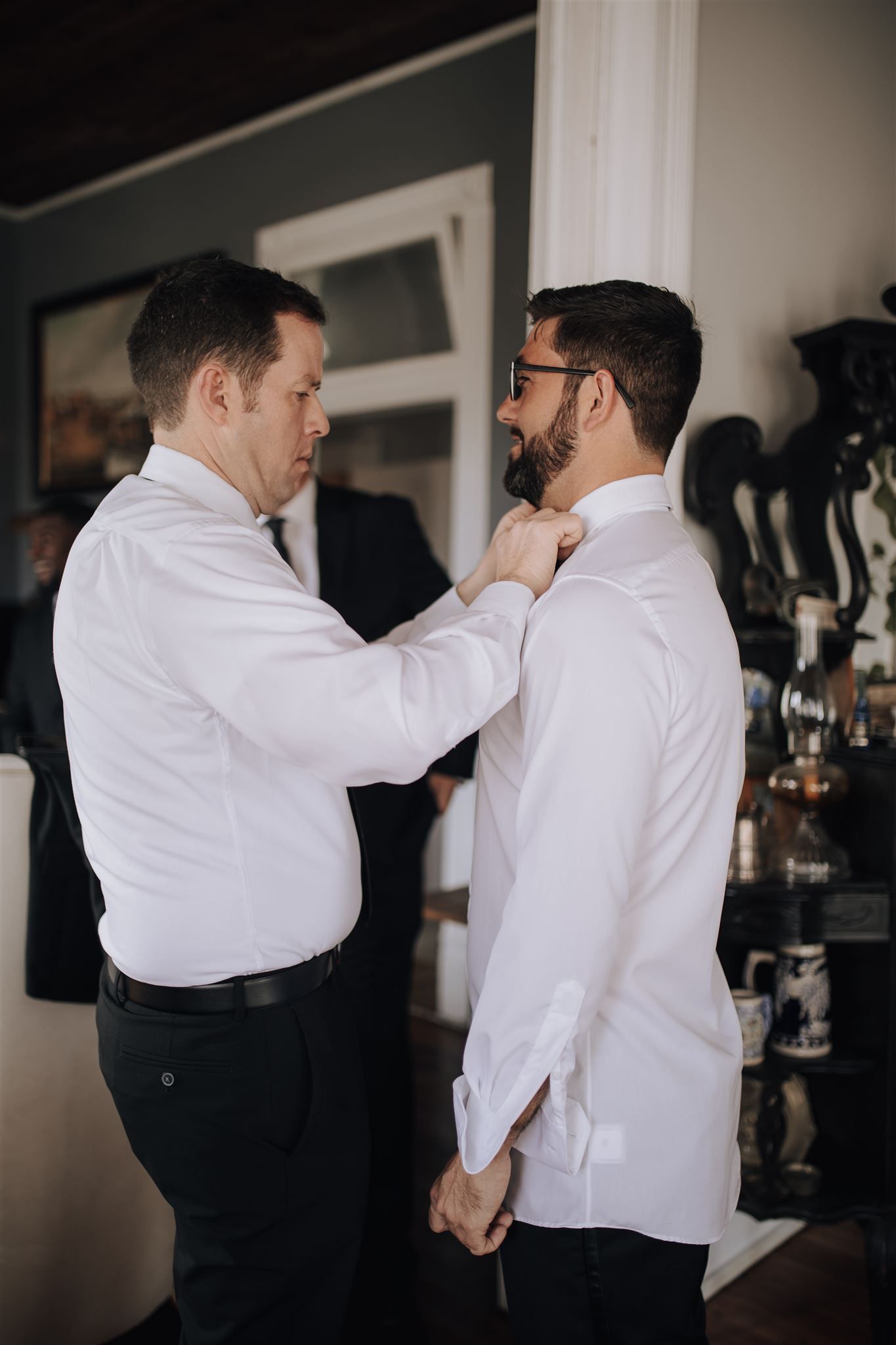 Two people in white dress shirts helping with wedding day preparations indoors.