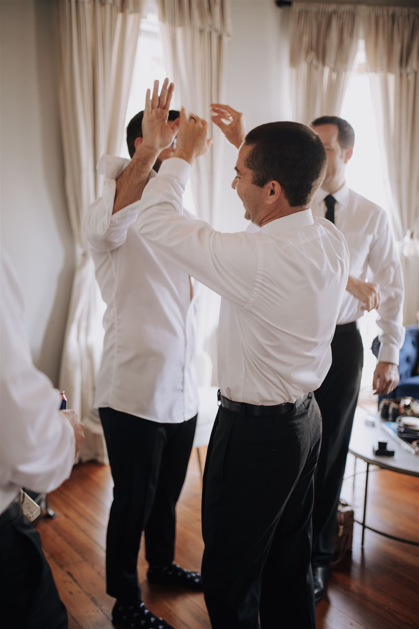 Groomsmen in white dress shirts prepare for a wedding celebration, helping each other get ready.