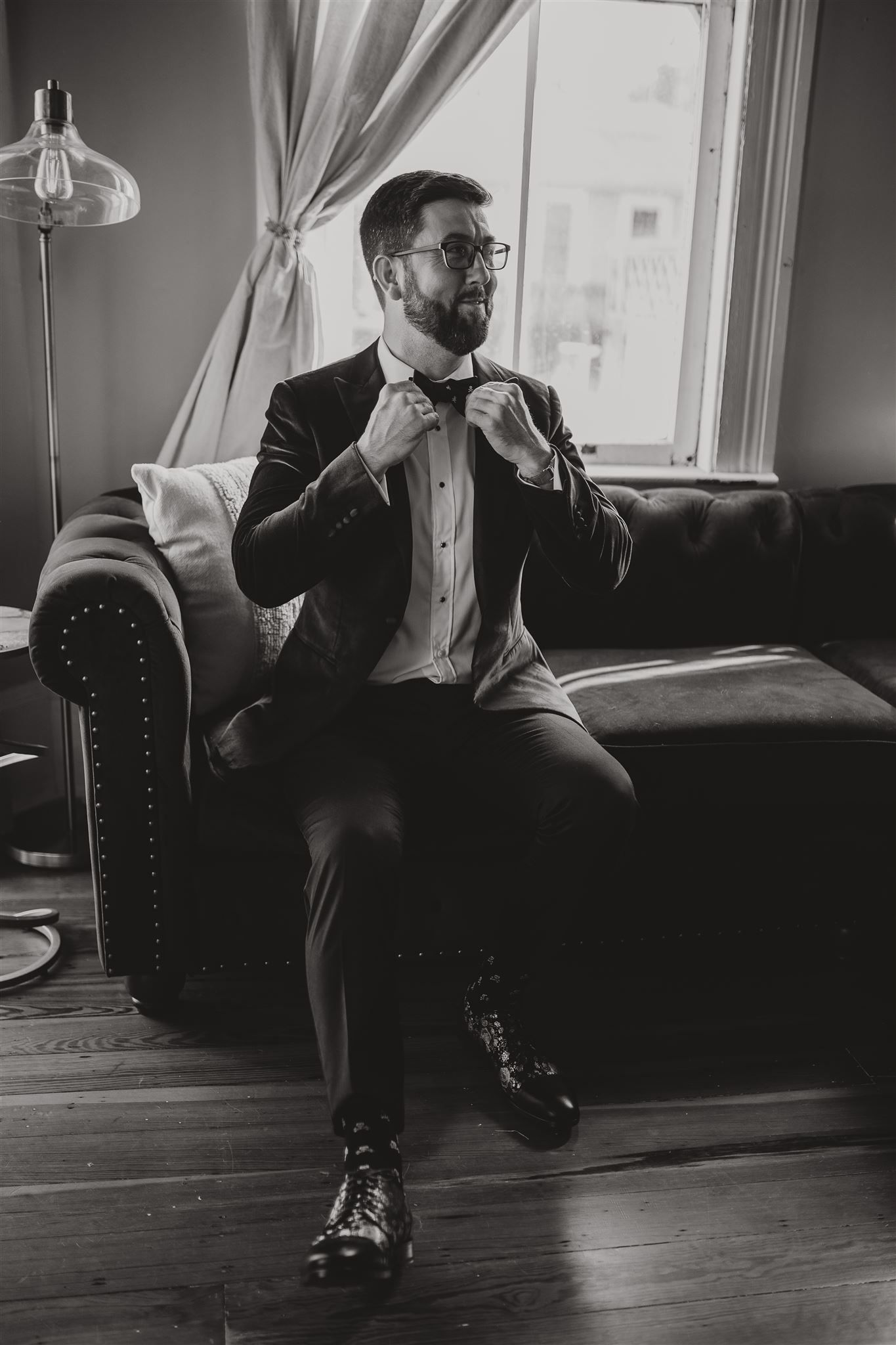 Person adjusting bowtie while seated on leather couch by window.