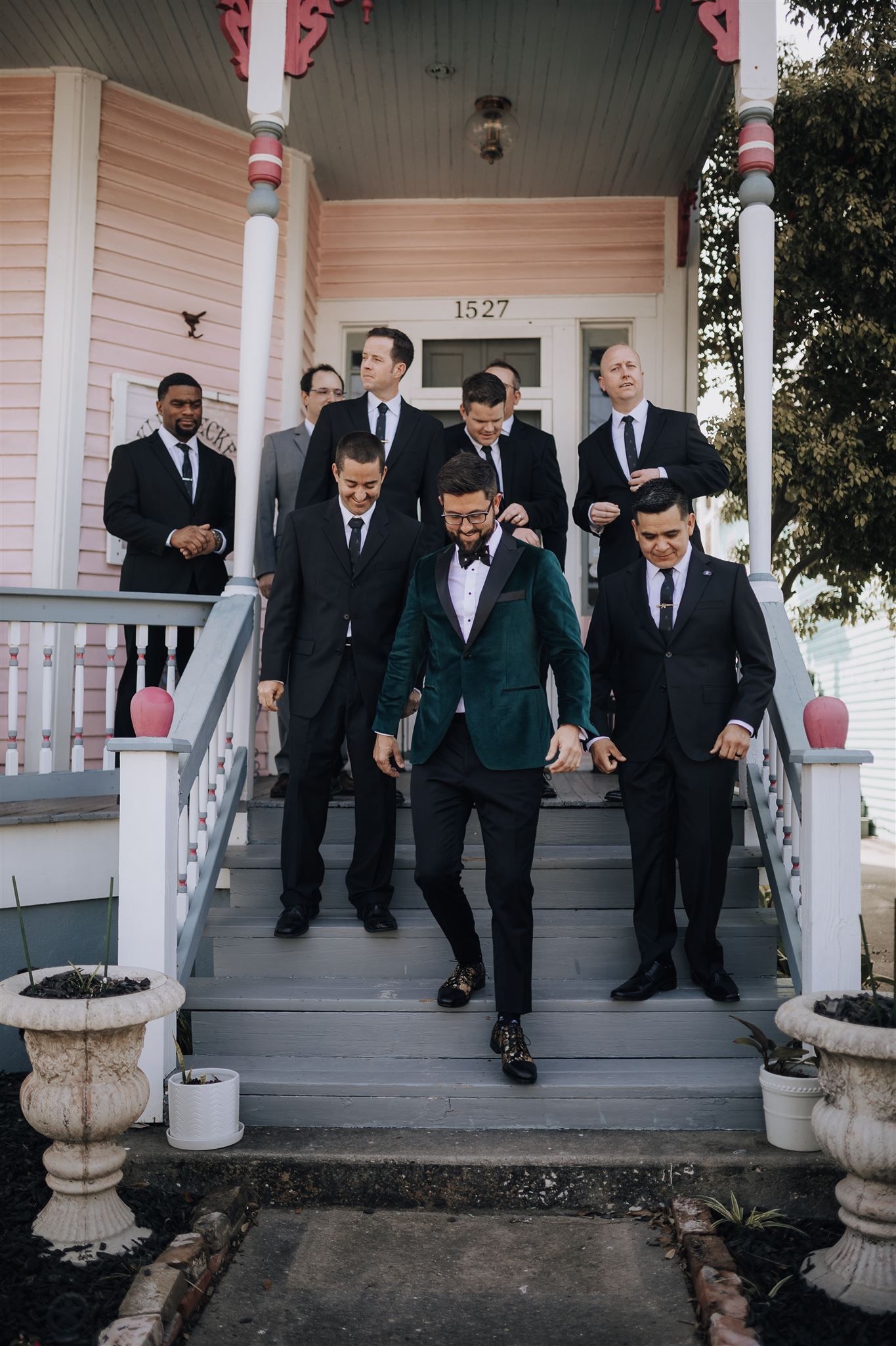 Wedding party in suits descending pink house stairs with groom in emerald green jacket.