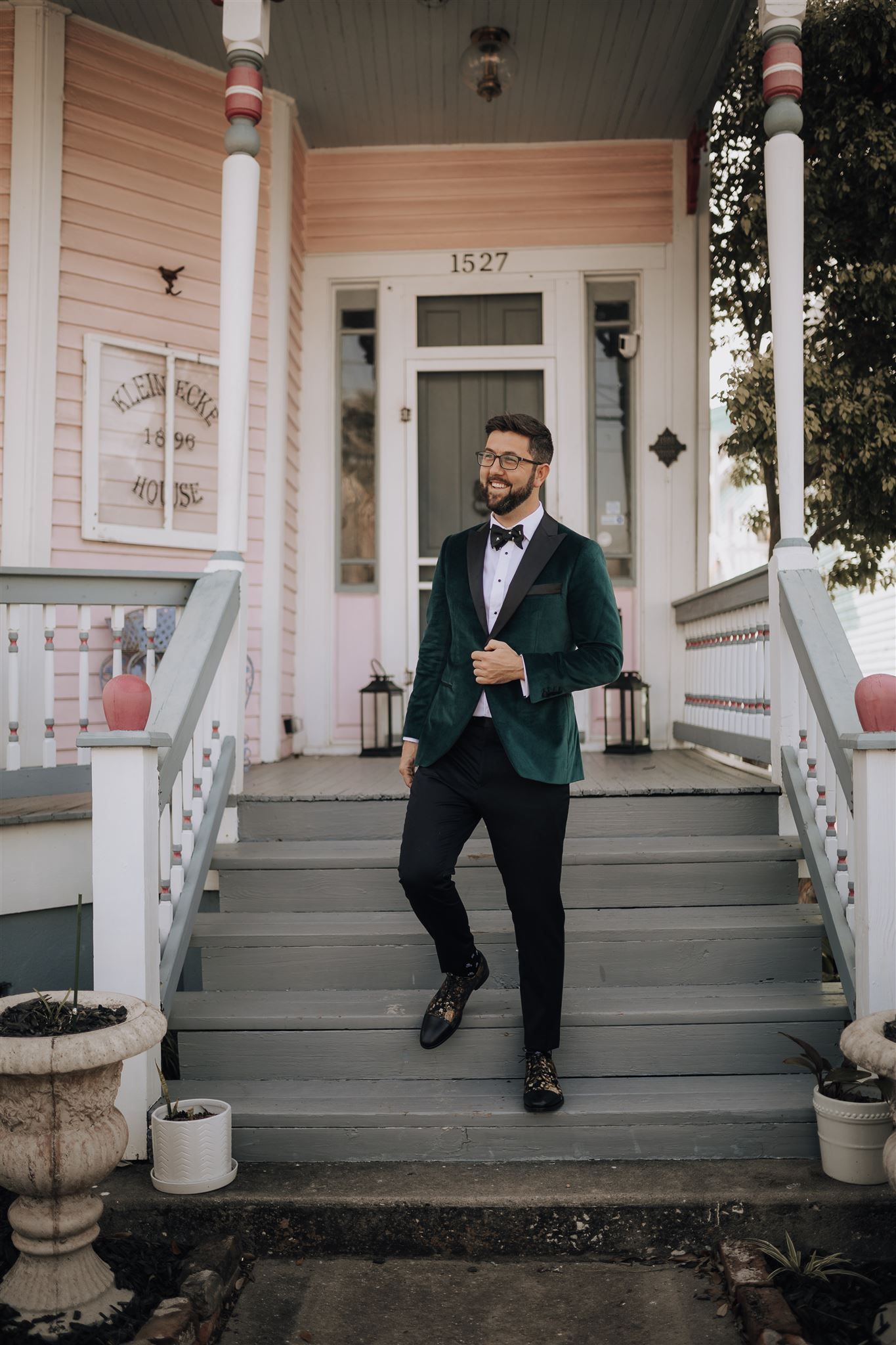 Person in emerald velvet tuxedo jacket poses on front porch of pink Victorian house.