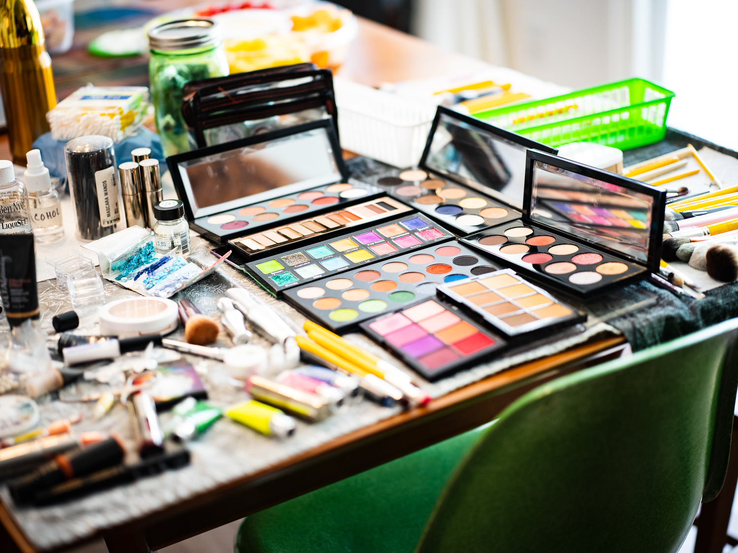 Professional makeup palette and tools scattered on green table with various cosmetic products.