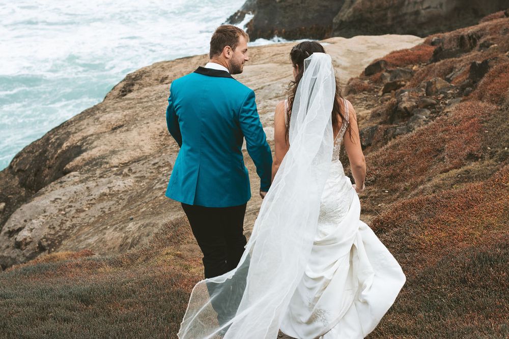 Couple in wedding attire stands on coastal cliffs overlooking ocean waves.
