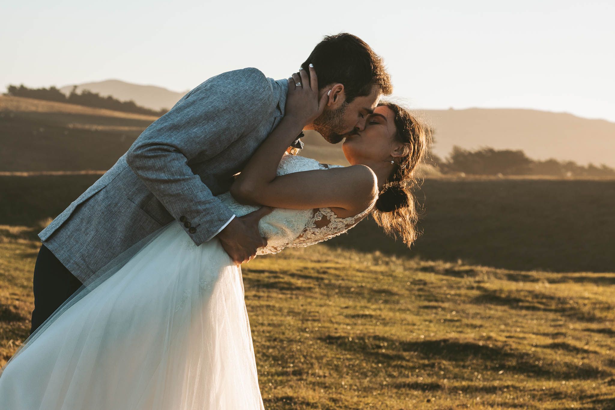 A romantic wedding couple shares a kiss during golden hour in a scenic hillside meadow.