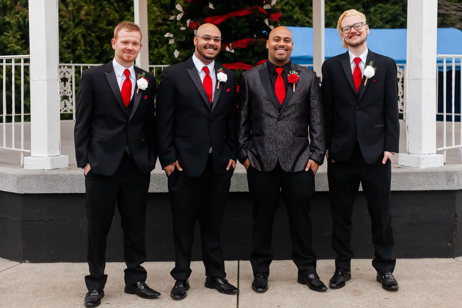 Groomsmen in black suits with red ties pose together outdoors.