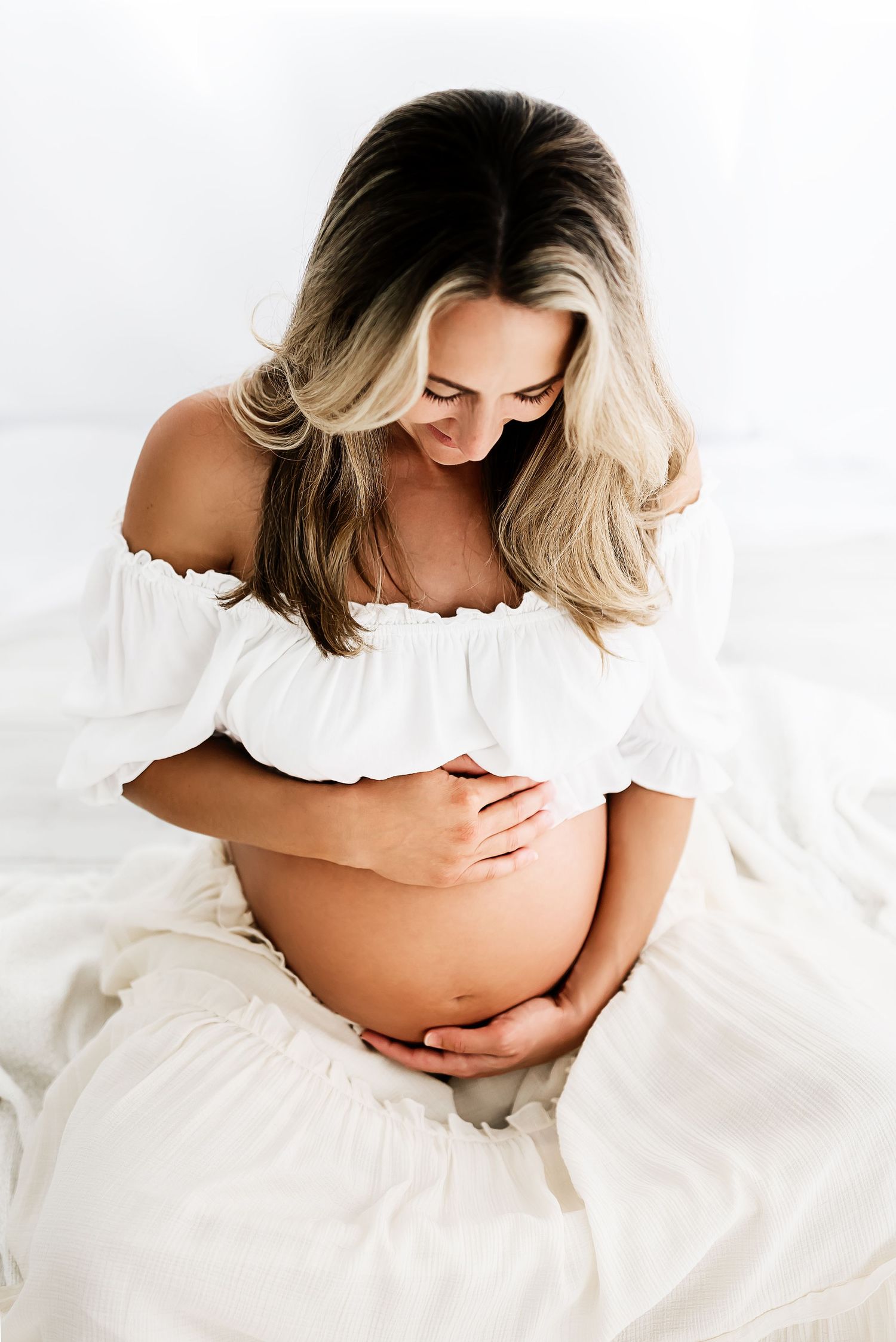 Maternity photos showing woman in white dress cradling her baby bump against light background.