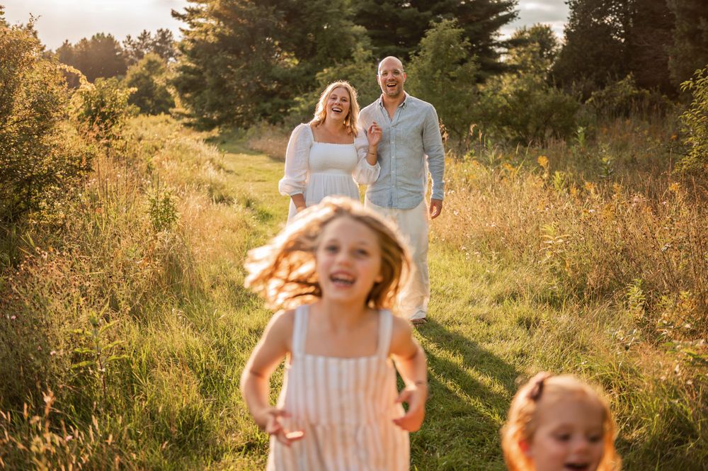 Children run joyfully down a sunlit path through tall grass while adults follow behind.