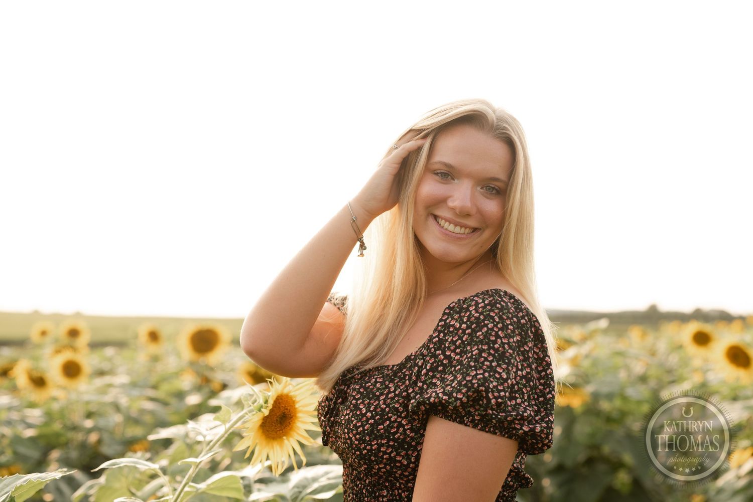 A casual summertime portrait with someone brushing hair back among sunflowers at sunset.