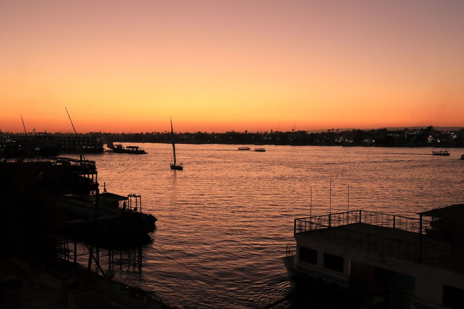 Silhouette of sailboats at sunset over calm harbor waters with orange sky.