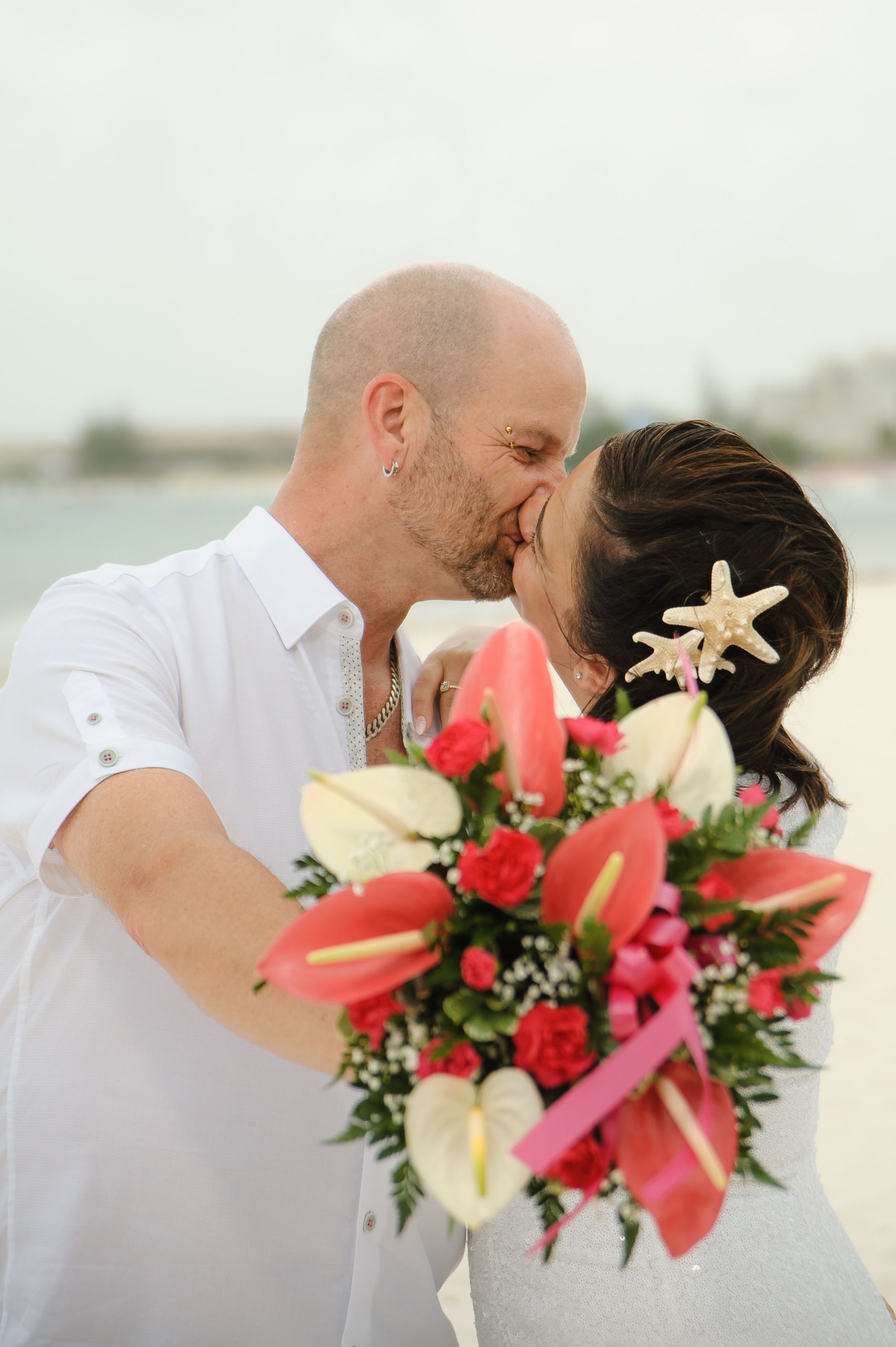 Wedding couple kisses while holding vibrant tropical bouquet near the water.