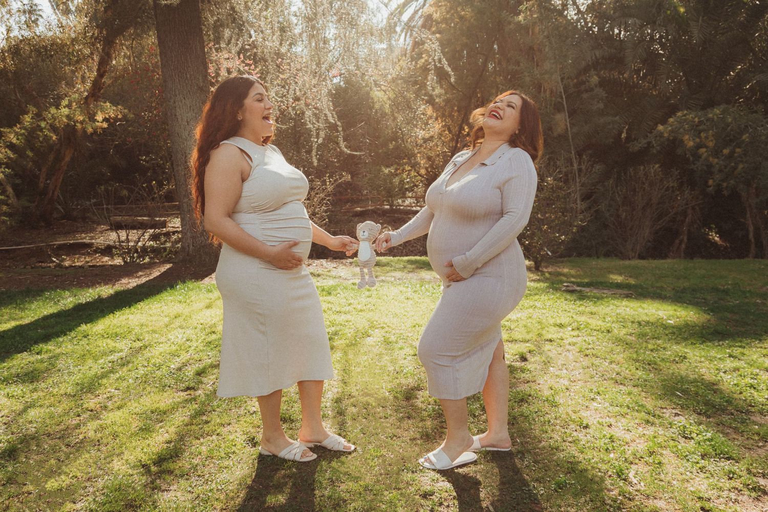 Two people in white dresses pose playfully in a sunlit garden setting with golden sunlight streaming through trees.