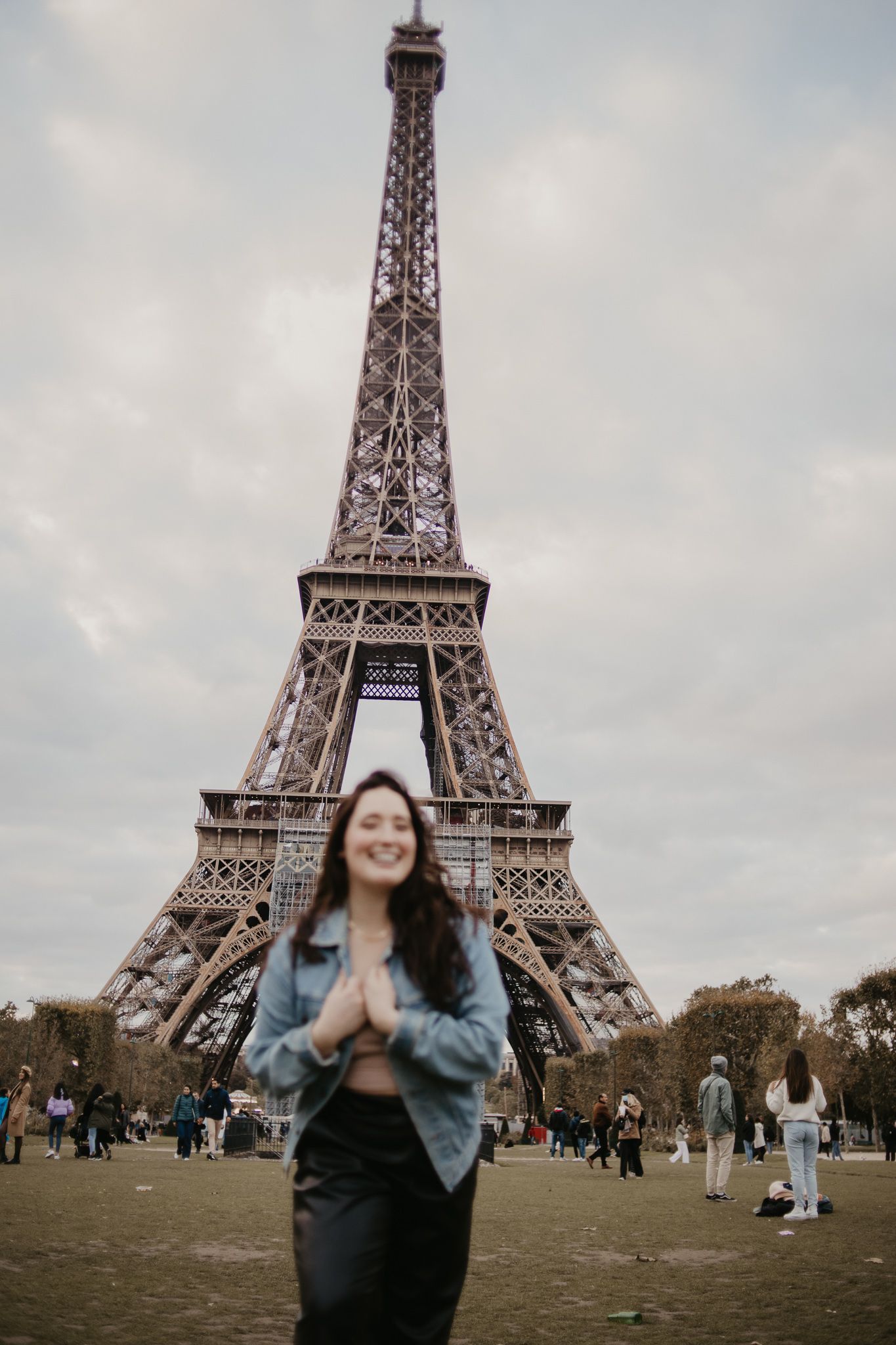 Tourist poses in front of the iconic Eiffel Tower in Paris on a cloudy day.