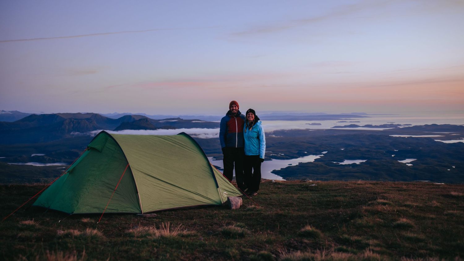 Green tent pitched on hilltop at sunset with mountains and lakes in background.