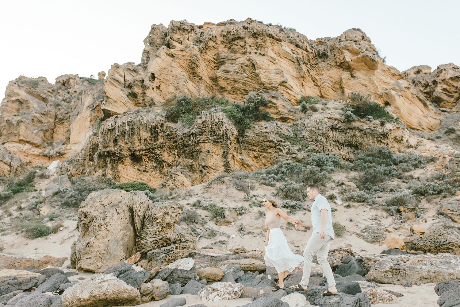 Couple exploring rocky coastal cliffs against a bright sky at sunset.